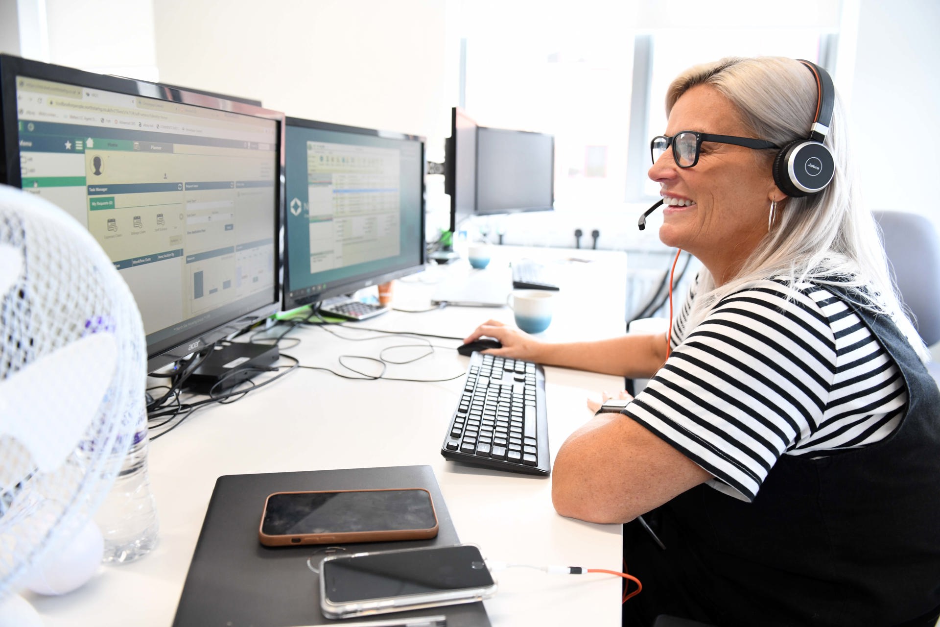  A smiling woman wearing a headset sits at a desk in a bright office, working on a computer with two large monitors. She is wearing glasses and a striped shirt, and appears to be in a customer service or support role. A water bottle, two smartphones, and a coffee mug are visible on the desk. The setting looks professional and friendly.